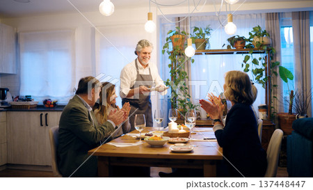 Senior man serving dinner to applauding friends at home Senior man serving dinner to applauding friends at home 137448447