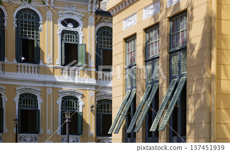 Golden sunlight spills across the creamy, light-hued facade of this Neoclassical structure, casting sharp shadows that accentuate the intricate scrollwork and deep textures of its architectural 137451939