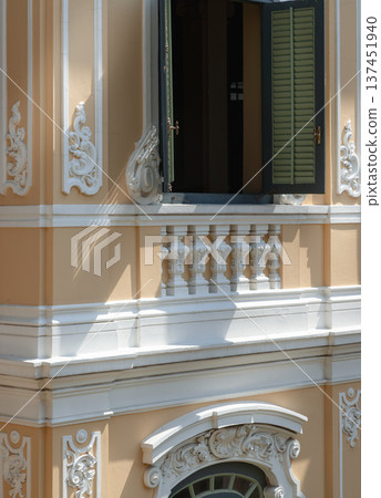 Sharp shadows play across the warm, light-colored facade, emphasizing the Neoclassical depth of the scrollwork. Amidst the architectural texture, an open window with dark green shutters stands as a Sharp shadows play across the warm, light-colored facade, emphasizing the Neoclassical depth of the scrollwork. Amidst the architectural texture, an open window with dark green shutters stands as a 137451940