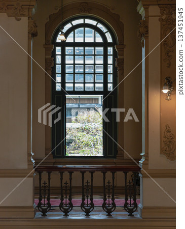 an elegant interior architectural scene featuring a large, tall, dark-framed arched window with a grid of smaller panes at its center. In the foreground, there is an intricately carved dark wooden or 137451945