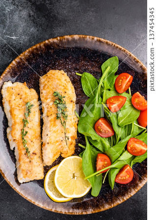Roast Cod fish fiilets, white fish meat on plate with salad. Black background. top view 137454345