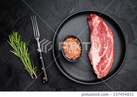 Raw beef chuck eye steak with rich marbling on black plate with rosemary and salt. Fresh beef cut overhead on dark background. Raw beef chuck eye steak with rich marbling on black plate with rosemary and salt. Fresh beef cut overhead on dark background. 137454831