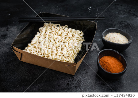 Dry instant ramen noodle block in black bowl with chopsticks and seasoning on dark background top view Asian convenience food concept Dry instant ramen noodle block in black bowl with chopsticks and seasoning on dark background top view Asian convenience food concept 137454920