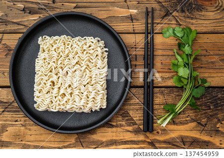 Dry instant ramen noodle block on black plate with chopsticks and fresh cilantro over rustic wooden background top view 137454959