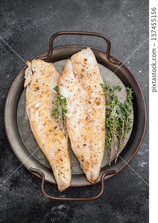 Baked cod style fish fillet, seasoned and herbed, served in iron pan on wooden board over dark textured backdrop. 137455156