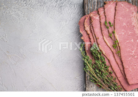 Thin pastrami slices with thyme on a rustic wooden serving board, placed over a textured gray background. Smoked deli beef ready for sandwiches or recipes. Thin pastrami slices with thyme on a rustic wooden serving board, placed over a textured gray background. Smoked deli beef ready for sandwiches or recipes. 137455501