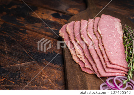 Smoked pastrami slices on a wooden board with a forged knife, red onion rings and fresh thyme. Premium deli beef presented on a dark rustic background, ideal for cooking or serving. Smoked pastrami slices on a wooden board with a forged knife, red onion rings and fresh thyme. Premium deli beef presented on a dark rustic background, ideal for cooking or serving. 137455543