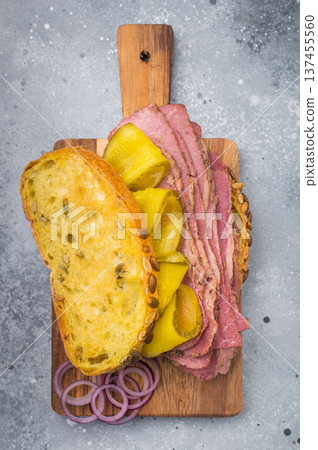 Gourmet pastrami sandwich with pickles and red onion on a wooden board over stone background, layered deli meat closeup. Gourmet pastrami sandwich with pickles and red onion on a wooden board over stone background, layered deli meat closeup. 137455560
