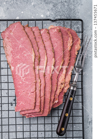 Smoked pastrami slices placed on a cooling rack next to a vintage fork on a gray stone surface. Premium deli beef ideal for sandwiches, serving or meal prep. 137455671