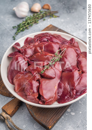 Raw turkey liver on a white plate, surrounded by garlic and thyme, set on a rustic wooden board against a neutral gray background. Raw turkey liver on a white plate, surrounded by garlic and thyme, set on a rustic wooden board against a neutral gray background. 137455680