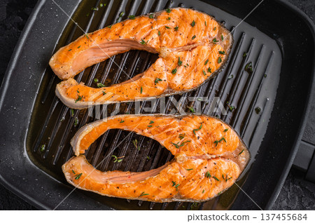 Two salmon steaks sizzle on a ribbed grill pan, with hot oil and herbs emphasizing the high-heat searing process during cooking. Two salmon steaks sizzle on a ribbed grill pan, with hot oil and herbs emphasizing the high-heat searing process during cooking. 137455684