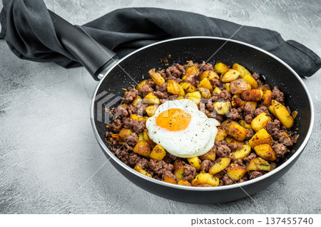 Beef hash with fried potatoes, ground beef and sunny egg in skillet on gray surface with dark napkin, perfect for rustic cooking themes. Beef hash with fried potatoes, ground beef and sunny egg in skillet on gray surface with dark napkin, perfect for rustic cooking themes. 137455740