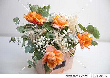 Close view of orange roses and delicate filler flowers arranged with eucalyptus in a pastel floral gift box. Romantic decoration, botanical elegance, festive surprise, and stylish flower arrangement Close view of orange roses and delicate filler flowers arranged with eucalyptus in a pastel floral gift box. Romantic decoration, botanical elegance, festive surprise, and stylish flower arrangement 137458397