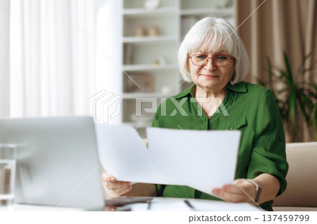 Senior woman reading documents while working on laptop at home 137459799