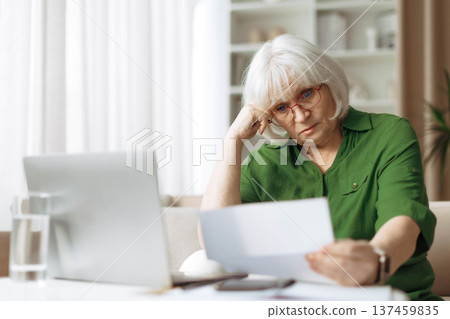 Senior woman looking worried while reading document at home Senior woman looking worried while reading document at home 137459835