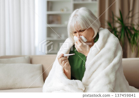 Senior woman checking body temperature with thermometer while wrapped in blanket 137459841