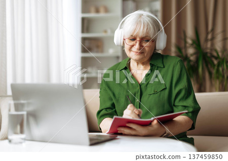 Senior woman writing notes in notebook and using laptop at home Senior woman writing notes in notebook and using laptop at home 137459850