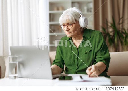 Senior woman wearing headphones while working on laptop at home desk Senior woman wearing headphones while working on laptop at home desk 137459851