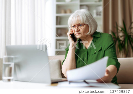 Serious senior woman reviewing document at home while speaking on phone Serious senior woman reviewing document at home while speaking on phone 137459857