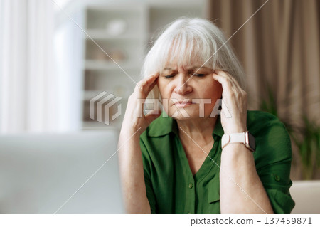 Senior woman suffering from headache while sitting at laptop at home Senior woman suffering from headache while sitting at laptop at home 137459871