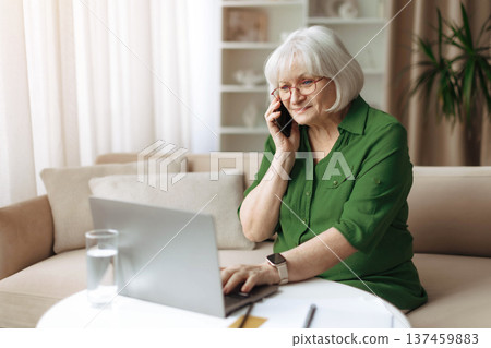 Senior woman talking on smartphone while working on laptop at home Senior woman talking on smartphone while working on laptop at home 137459883