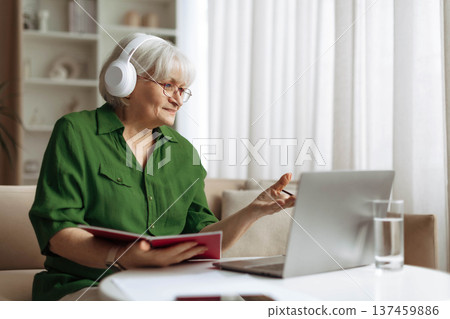 Senior woman wearing headphones holding notebook while communicating through laptop at home Senior woman wearing headphones holding notebook while communicating through laptop at home 137459886