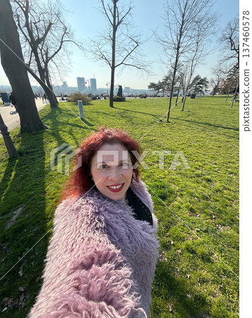 Smiling woman taking selfie in sunny park with green grass and trees. Outdoor lifestyle, spring atmosphere and joyful urban recreation moment. 137460578