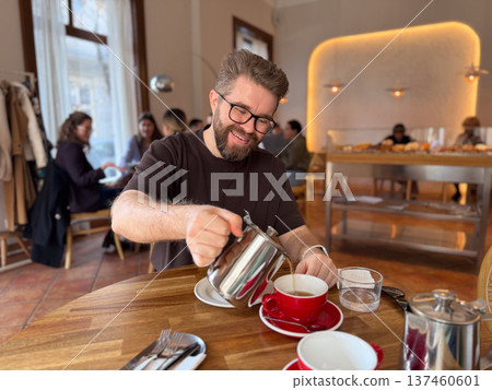 Man pouring tea from metal teapot into red cup while sitting at cafe table. Relaxed breakfast scene, urban cafe culture and casual morning lifestyle. 137460601