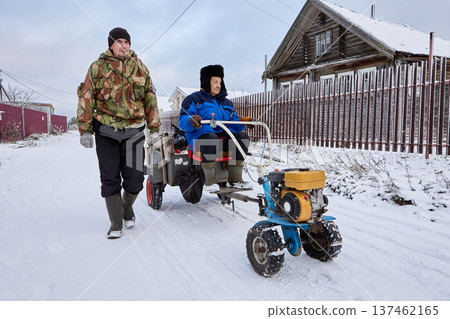 Person operates motorized unit on snowy village street, companion walks beside, wooden houses and bare trees in background. 137462165