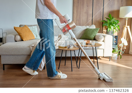 Woman vacuuming floor with cordless vacuum cleaner in living room with sofa and tables 137462326