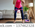 Woman doing squat exercise with resistance band on mat, with dumbbell and water bottle nearby 137462328
