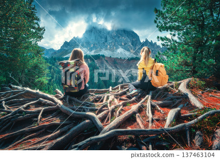 Two young women with backpacks sitting on tree roots in mountain Two young women with backpacks sitting on tree roots in mountain 137463104
