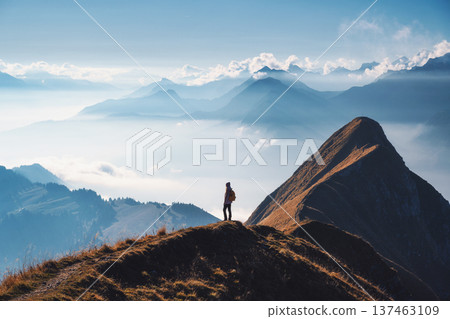 Woman with backpack standing on a mountain ridge above clouds Woman with backpack standing on a mountain ridge above clouds 137463109