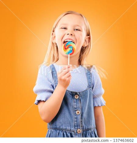 Lollipop, girl child and portrait of candy in hand, eating and studio for sweets, birthday party or carnival. Sugar, kid and lick spiral snack for dessert, hunger or meal on orange background 137463602