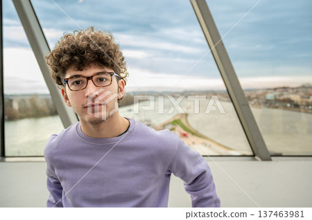Young man with curly hair and glasses wearing a purple sweater, looking directly at the camera with a blurred panoramic city and river view behind him Young man with curly hair and glasses wearing a purple sweater, looking directly at the camera with a blurred panoramic city and river view behind him 137463981