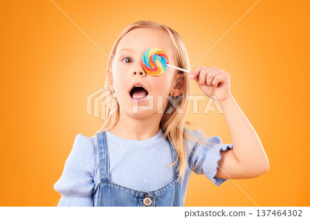 Portrait, cute and a child with a lollipop on a studio background for food, candy or crazy expression. Orange, comic and a girl kid with sweets, treat or sugar for covering an eye on a backdrop 137464302