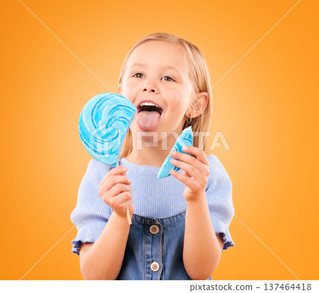 Child, eating lollipop or candy in studio for sweet tooth, color spiral or sugar for energy. Face of happy girl kid on orange background for snack, treat or thinking of dessert or unhealthy food 137464418