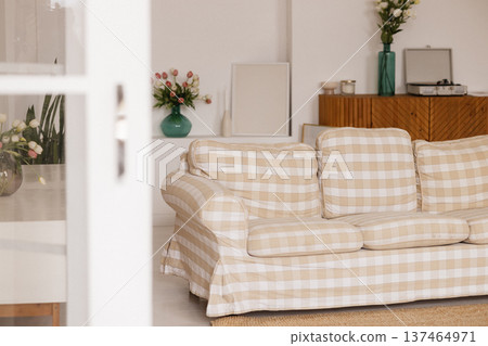 Beige plaid sofa standing in living room with white wall, wooden cabinet and flowers 137464971