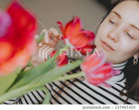 Young woman admiring red tulips bouquet at home. Gentle aesthetic of spring morning and self care. Blurred flowers in foreground. Concept of mindfulness, seasonal joy, and natural female beauty. 137465305