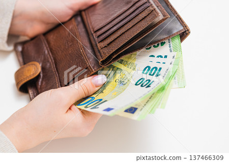 Close up of woman placing 100 euro banknotes into brown leather wallet. Concept of savings, personal finance, cash payment, budgeting, wealth and financial security in Europe. Close up of woman placing 100 euro banknotes into brown leather wallet. Concept of savings, personal finance, cash payment, budgeting, wealth and financial security in Europe. 137466309