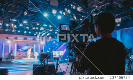 A camera operator captures a stage performance in a TV studio. Bright lights illuminate the set as crew members prepare for the live broadcast. Cameras are set up for different angles. 137466724