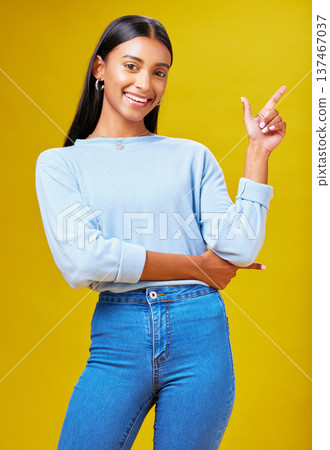 Pointing, mockup and portrait of a woman in a studio for advertising, marketing or promotion. Smile, happy and young Indian female model with a direction hand gesture isolated by yellow background. Pointing, mockup and portrait of a woman in a studio for advertising, marketing or promotion. Smile, happy and young Indian female model with a direction hand gesture isolated by yellow background. 137467037