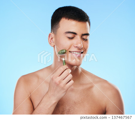 Man, beauty and face with skincare, roller for skin and grooming with smile on blue background. Jade crystal, model and dermatology, cosmetic tools and treatment with facial massage in a studio 137467567