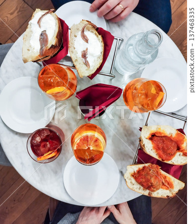 Overhead view of an italian aperitivo table featuring refreshing spritz cocktails, a dark red drink, water, and various appetizers including anchovy and meatball sandwiches Overhead view of an italian aperitivo table featuring refreshing spritz cocktails, a dark red drink, water, and various appetizers including anchovy and meatball sandwiches 137468335