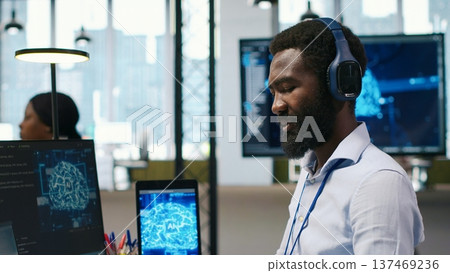 Computer scientist in office integrating AI machine learning systems to streamline workflows, earing headphones. African american man uses artificial intelligence on devices, listening music, camera A Computer scientist in office integrating AI machine learning systems to streamline workflows, earing headphones. African american man uses artificial intelligence on devices, listening music, camera A 137469236