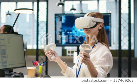 Female technician wearing VR goggles coding with AI language model assistance in startup office. Young woman using virtual reality integrating LLM assistant suggestions into code editor, camera A 137469262