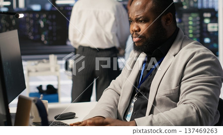 Stressed technician in data center managing AI server infrastructure, drinking coffee. African american worker using PC to run artificial intelligence, panicked by deadline, camera A 137469263