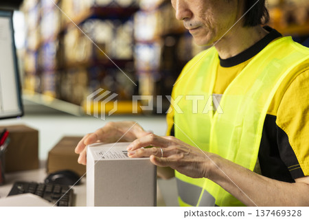 Logistics center employee manages packaging tasks and labeling, applying printed airway bill tracking number on packages. Man in hi vis vest packing orders in storage room depot. Logistics center employee manages packaging tasks and labeling, applying printed airway bill tracking number on packages. Man in hi vis vest packing orders in storage room depot. 137469328