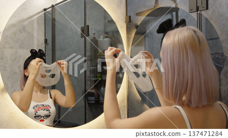 Woman applying sheet mask on face in front of mirror Woman applying sheet mask on face in front of mirror 137471284