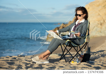 Businesswoman sits on a beach chair with a laptop and whiskey 137474386